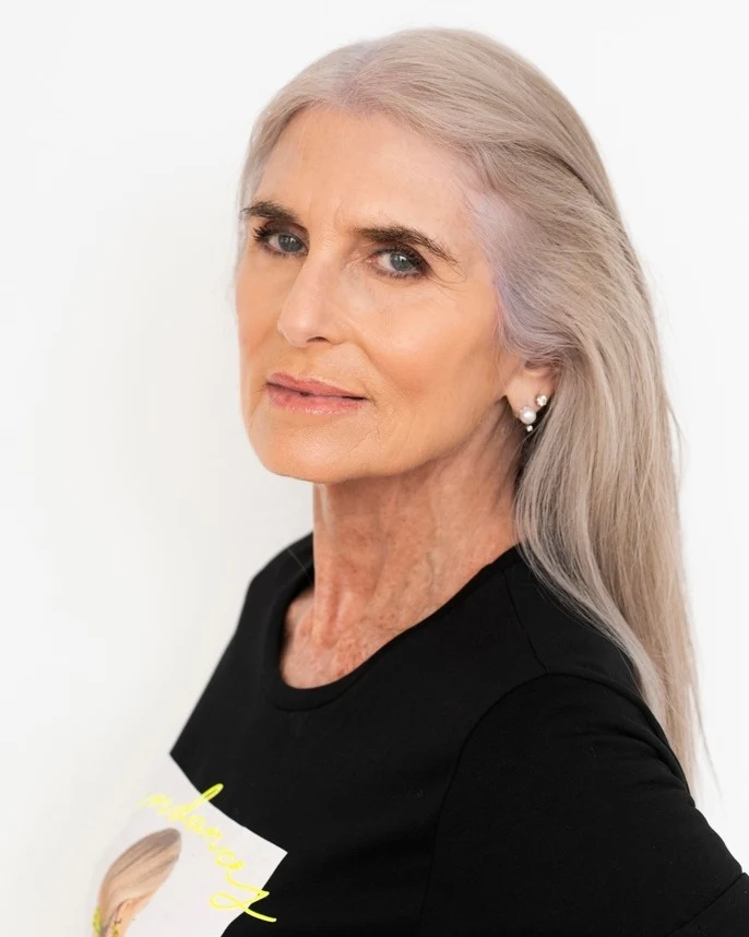 A woman over 60 with long straight silver hair styled with a center part and tucked behind her ears.