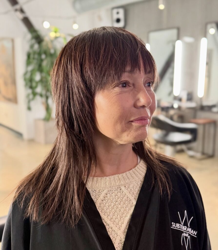A woman over 60 inside a salon showing a mid-length brunette shag hairstyle with choppy layers and full straight bangs.
