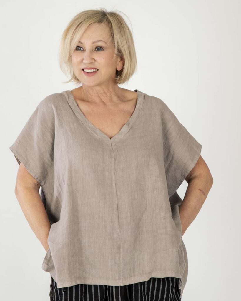 A smiling woman with a shoulder-length layered ash blonde lob haircut and wispy curtain bangs wearing a black patterned top.