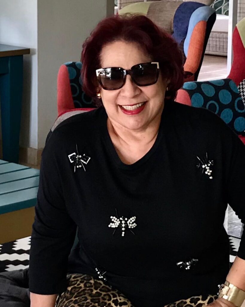 A smiling woman with a short voluminous mahogany red hairstyle wearing sunglasses and a black top.