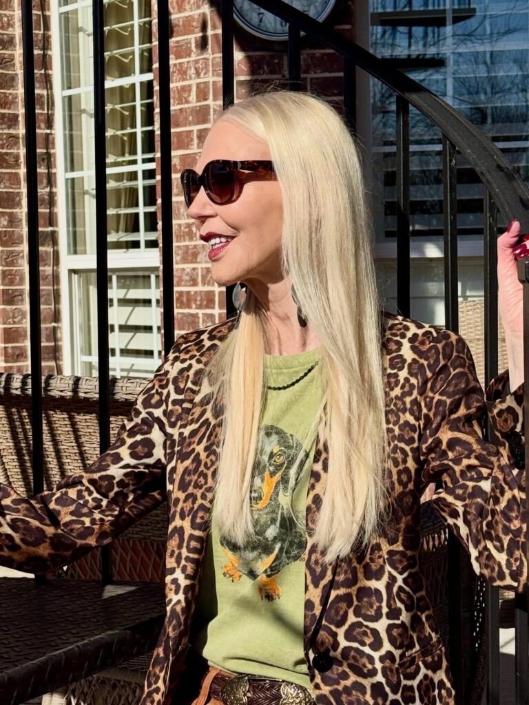 A smiling woman with long straight honey blonde hair and sunglasses posing outdoors on a walkway.