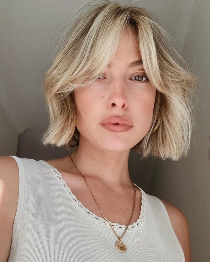 A woman with a short blonde blunt bob styled with natural waves and parted curtain bangs that frame her face.