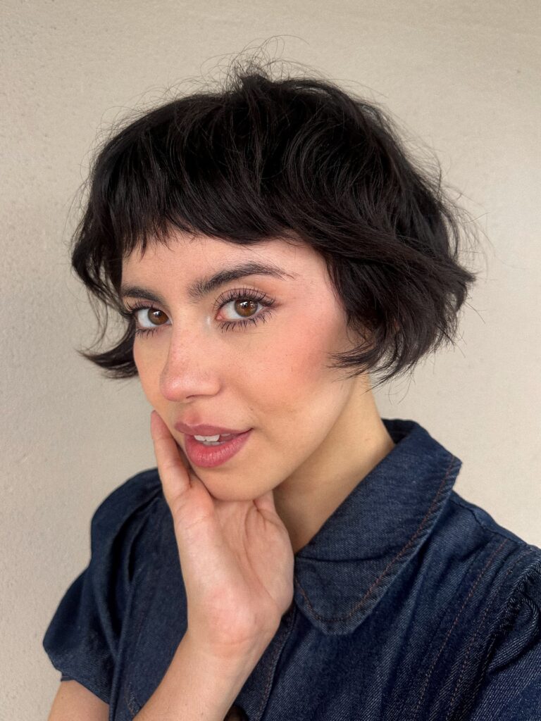 Portrait of a woman with dark wavy bixie cut hair and micro bangs.