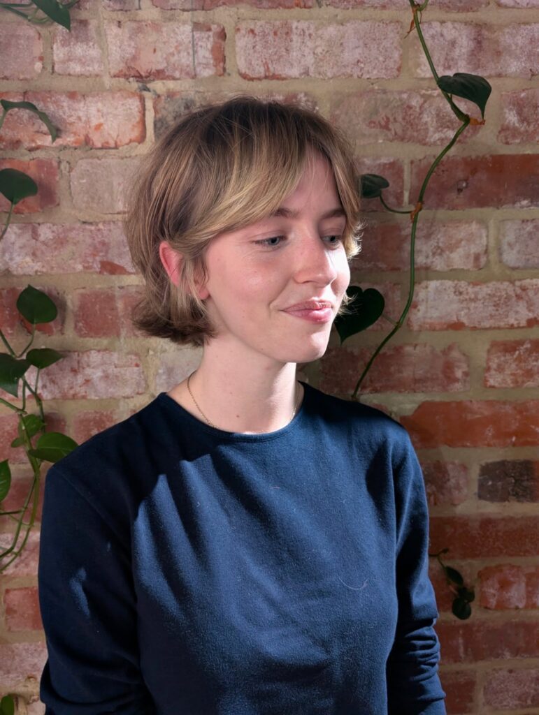 A young woman smiling in front of a brick wall showcasing a short honey blonde bixie cut with parted curtain bangs.