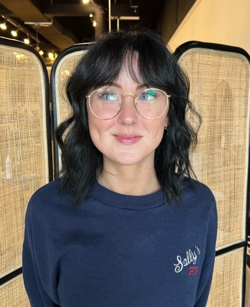 A portrait of a smiling woman with glasses showing off a textured jet black shoulder length shag haircut with curtain bangs.
