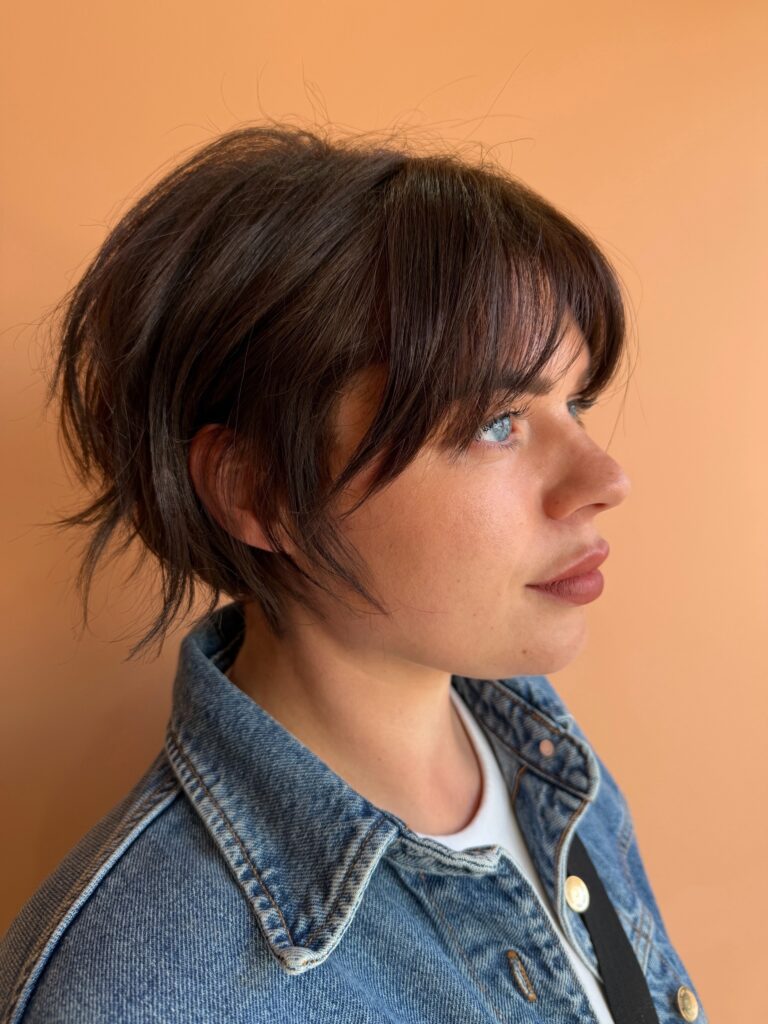 A young woman with a short, dark brunette pixie bob haircut featuring choppy layers and wispy, see-through bangs.