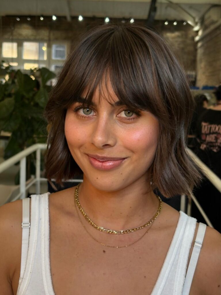 A smiling woman with green eyes showing off a short brunette wavy bob haircut with soft wispy bangs.