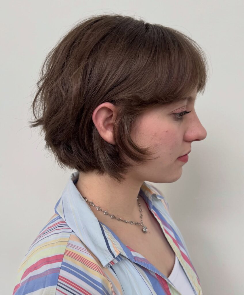 Side profile view of a woman with a cool ash brown bixie haircut featuring curved face framing tendrils and soft rounded bangs.