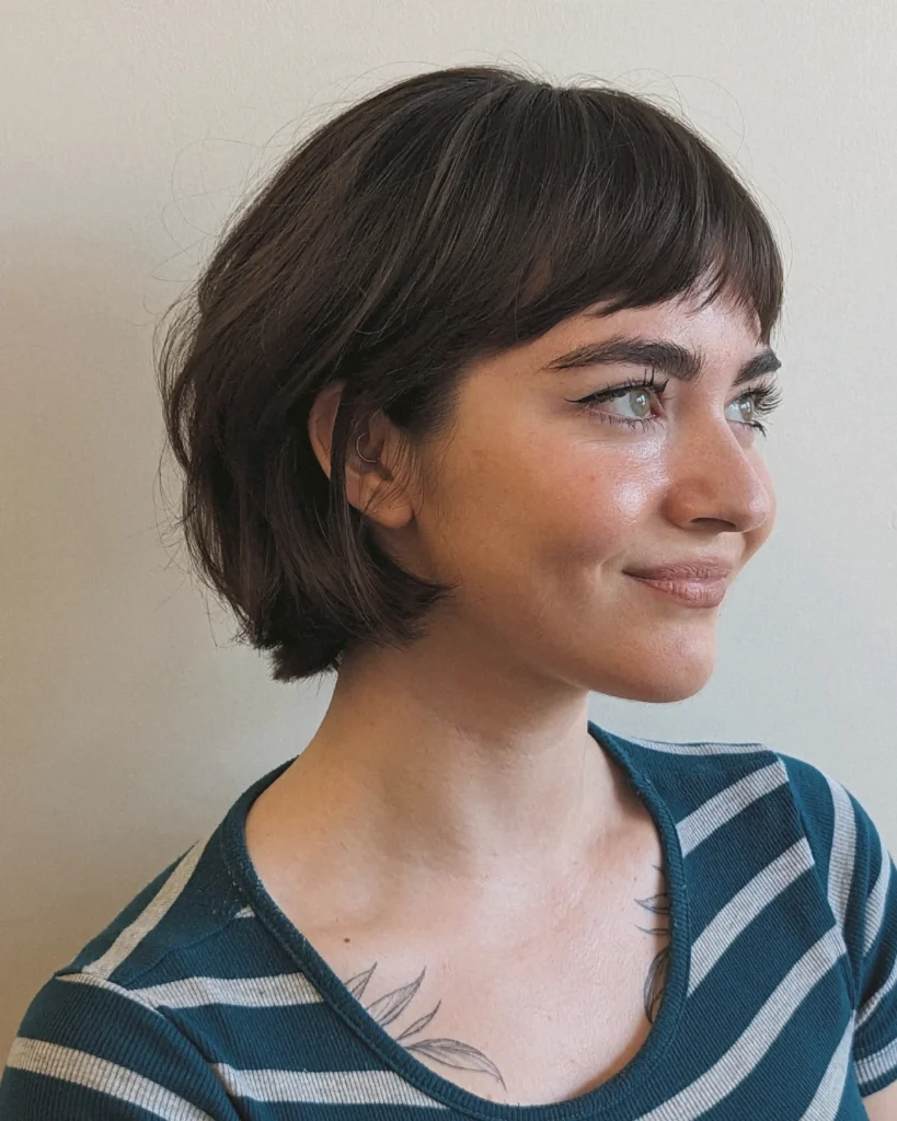 Side profile of a woman smiling with a short dark brown French bob tucked behind her ear and micro baby bangs.