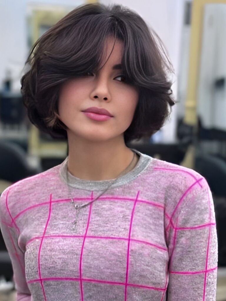 A portrait of a woman wearing a pink sweater with a short dark brown voluminous bob and sweeping curtain bangs.