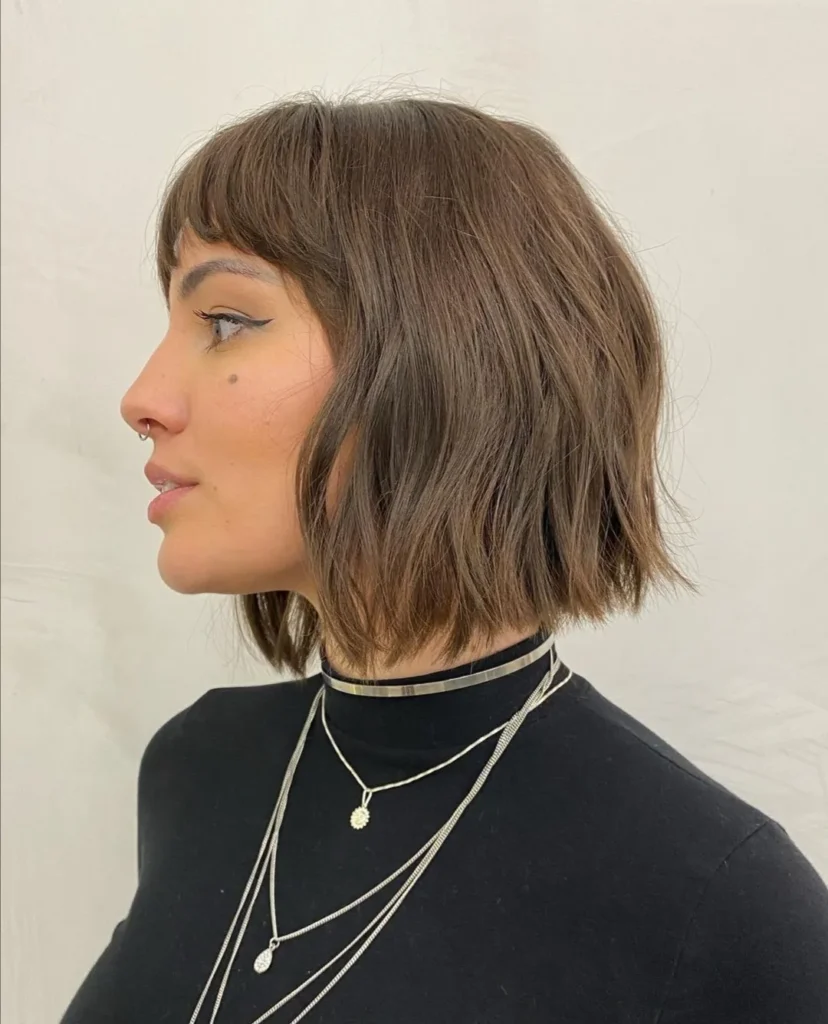 Side profile view of a woman with a chin length textured brown bob and full, straight-across blunt bangs.