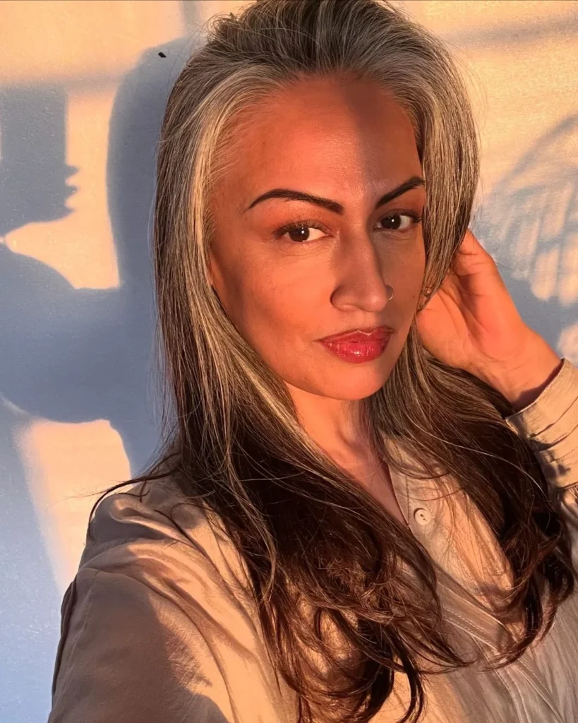 A woman with long wavy brunette hair and a striking silver money piece across her front hairline.