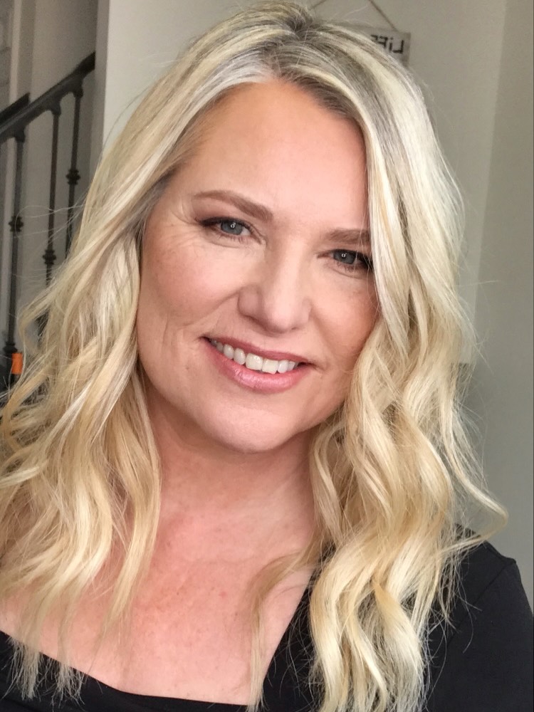 A smiling woman with long platinum blonde hair styled in beach waves with natural silver roots.
