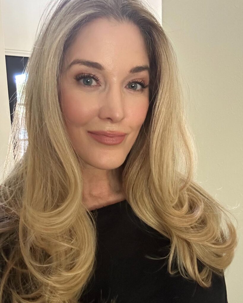 A close-up photograph of a smiling woman with long, voluminous, bouncy blonde hair styled in a center part.
