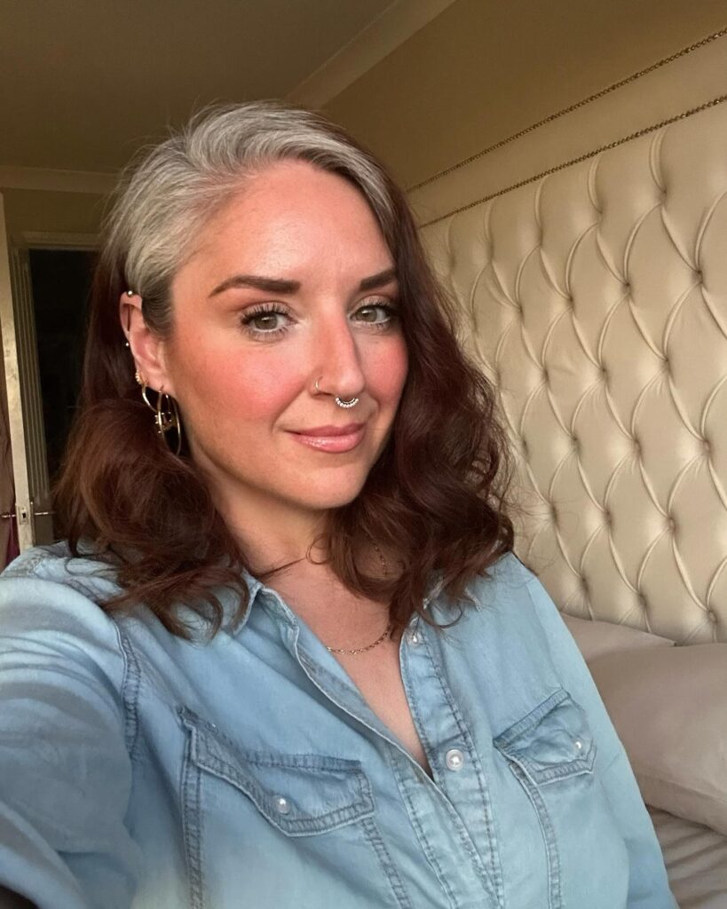 A woman with medium length wavy auburn brown hair featuring a prominent natural silver streak along her hairline.
