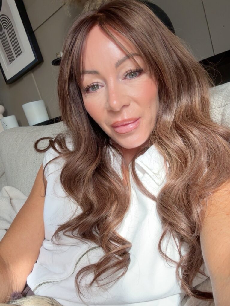 A woman with long wavy chestnut brown hair and soft curtain bangs smiling while sitting on a couch.