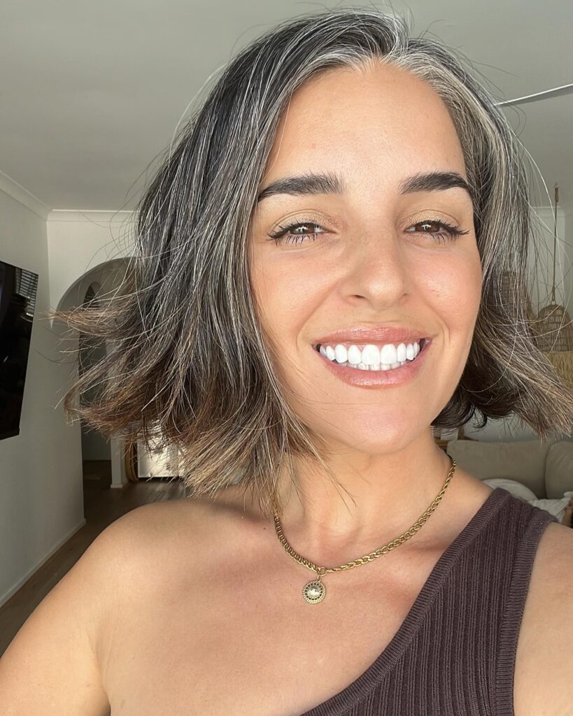 A smiling woman with a wavy jaw length salt and pepper bob haircut with silver highlights.