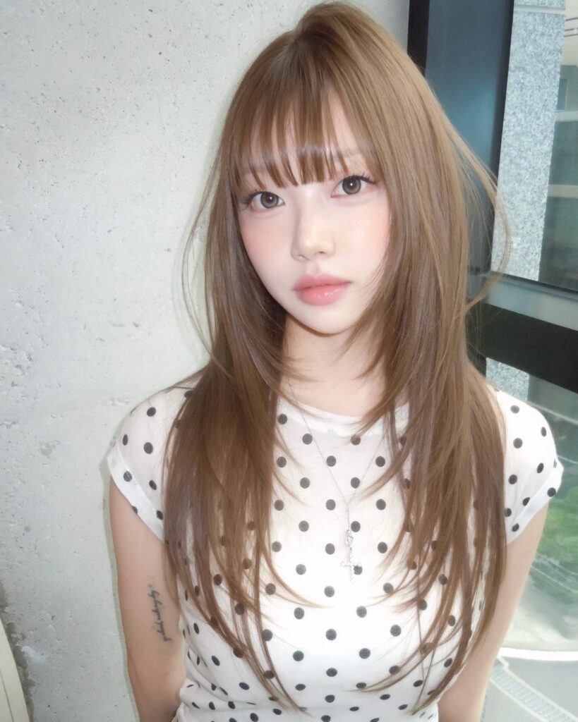 A young woman wearing a polka dot top poses with long light ash brown layered hair and see through bangs.
