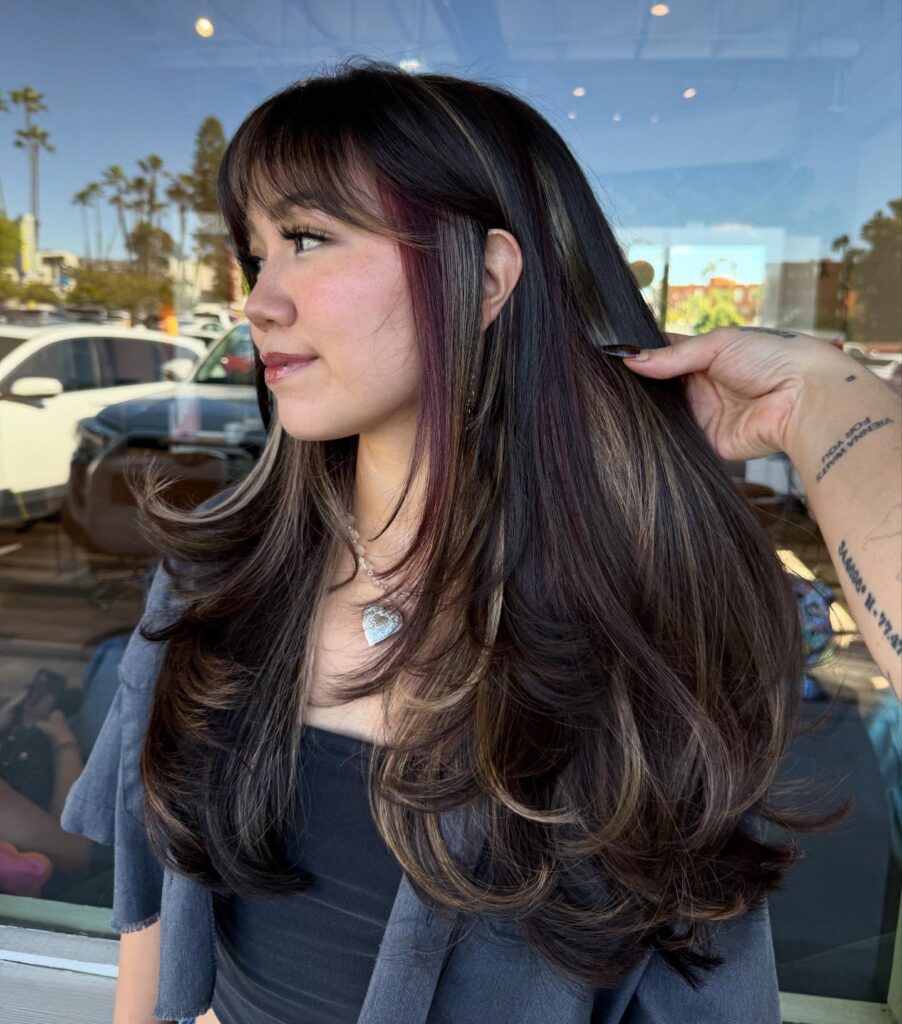 A young woman with very long layered espresso brown hair featuring hidden face framing pink and light brown highlights, styled with large bouncy waves and subtle bangs.