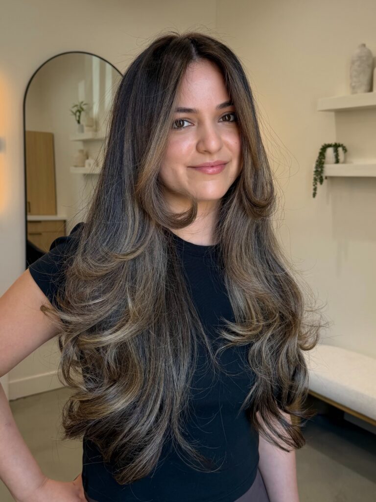 A smiling woman with long dark brown hair featuring ash bronde highlights and bouncy face framing layers.