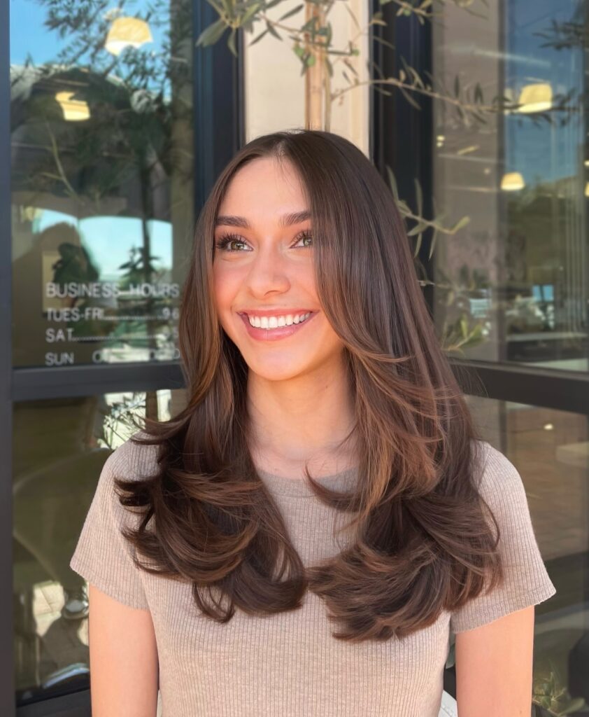 A smiling young woman with long glossy chocolate brown hair featuring soft face framing layers styled in a bouncy blowout.