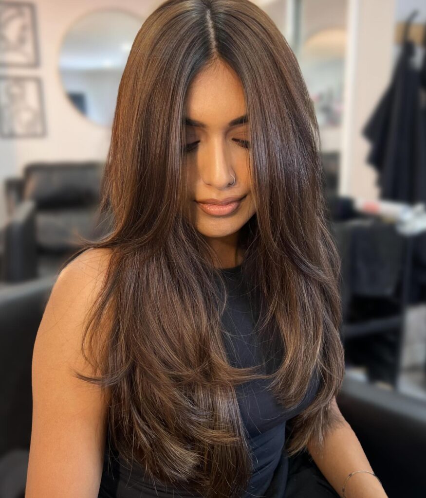 A woman with long, shiny chestnut brown hair with subtle warm highlights and soft layers looking down.