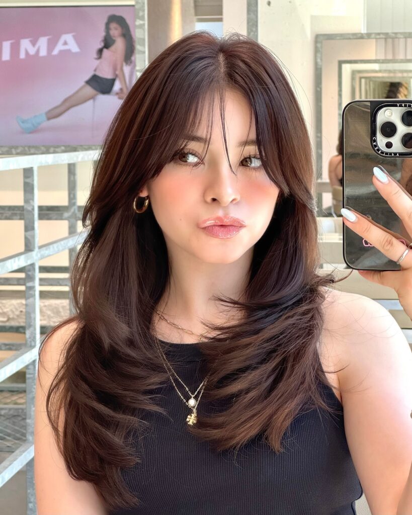 A woman with long layered dark brown hair and curtain bangs takes a mirror selfie, showcasing a soft, voluminous hairstyle with face-framing layers.