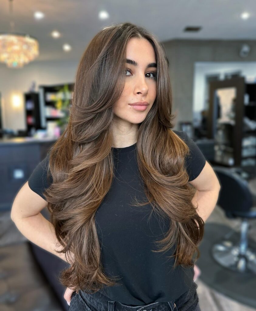 A woman with very long, thick chocolate brown hair featuring subtle caramel highlights, large voluminous waves, and swept back face framing layers, posed in a salon.