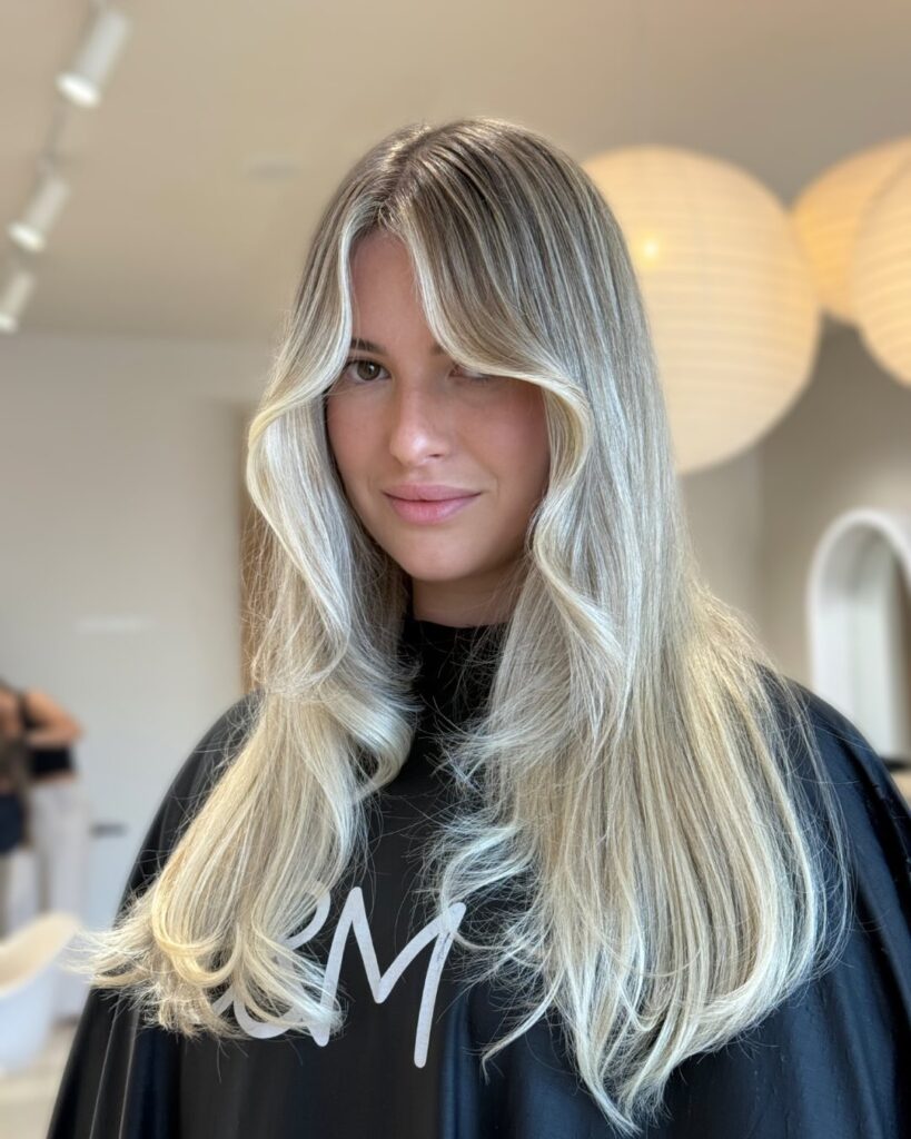 A young woman with long ash blonde layered hair featuring bright face framing curtain bangs and softly flipped ends.