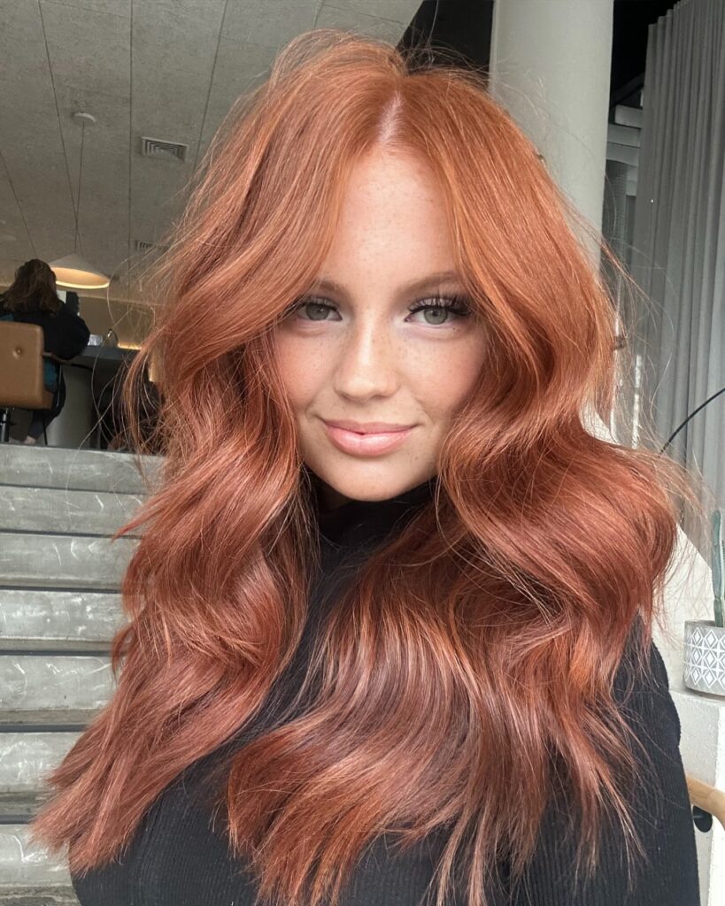 A young woman with long vibrant copper red hair styled in large beach waves poses on some steps.