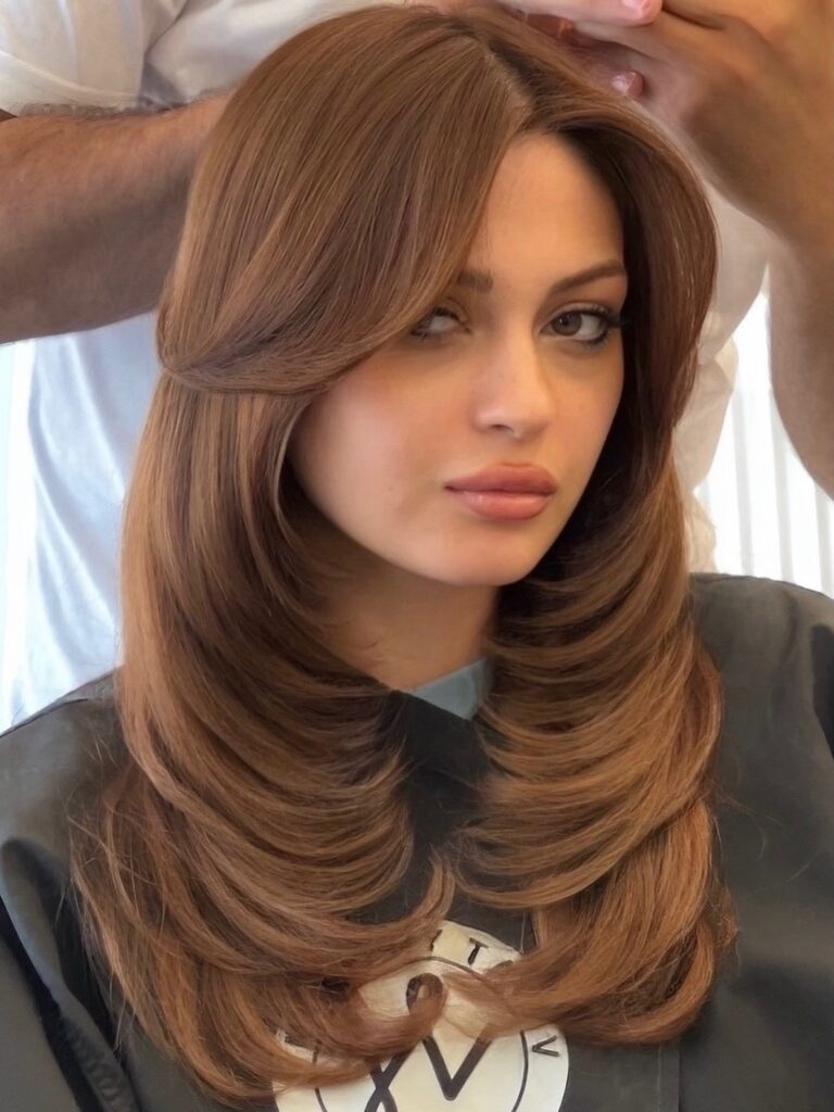 A young woman with medium length caramel chestnut brown hair styled in soft inward curved layers with a blowout finish.