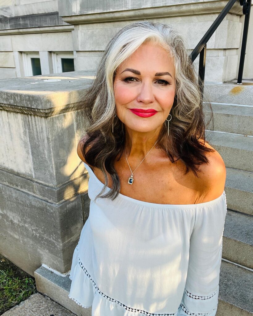 A woman with shoulder-length wavy hair displaying a prominent silver-white money piece strand against darker brunette hair.