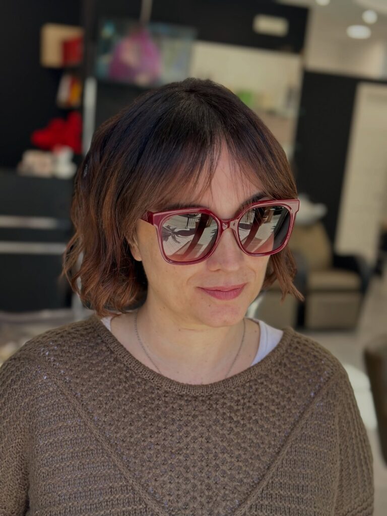 A woman wearing oversized red sunglasses with a chin-length choppy chestnut bob featuring soft waves and wispy see-through bangs.