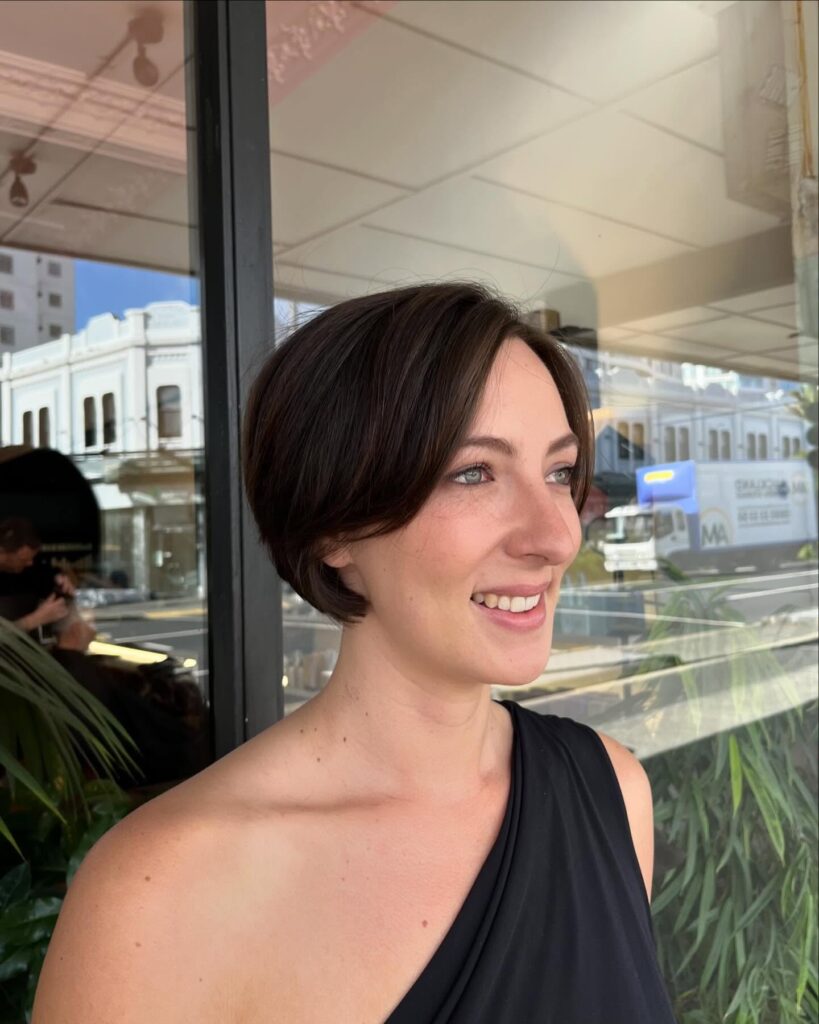 A woman smiling with a sleek, short espresso brown choppy bob featuring delicate face-framing curtain bangs.