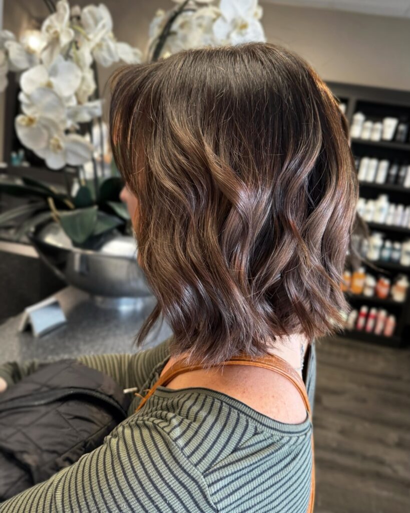 A side profile view of a woman with a choppy, chin-length espresso brunette bob featuring subtle caramel teasylights and loose waves.