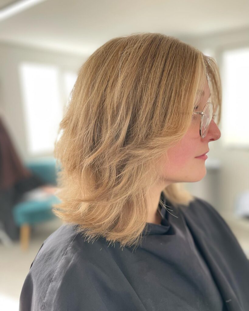A woman in profile wearing glasses showcases a honey blonde shoulder-length choppy bob with feathered face-framing layers.