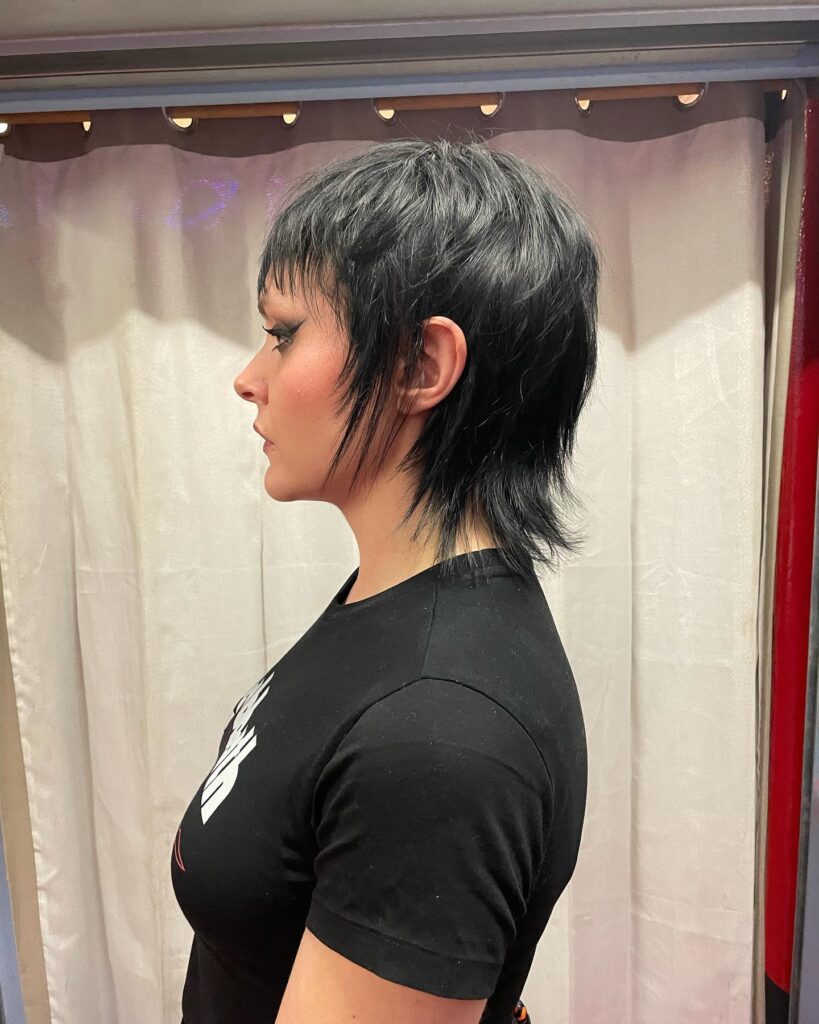 A woman in profile displays an edgy jet black shaggy mullet bob with a short choppy fringe and long feathered layers at the back.