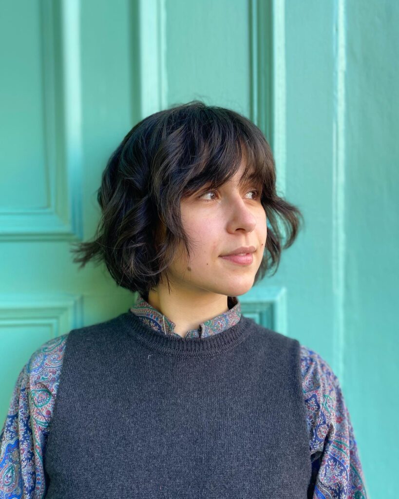 A woman with a dark brunette choppy chin-length bob featuring soft waves and wispy curtain bangs poses against a green door.