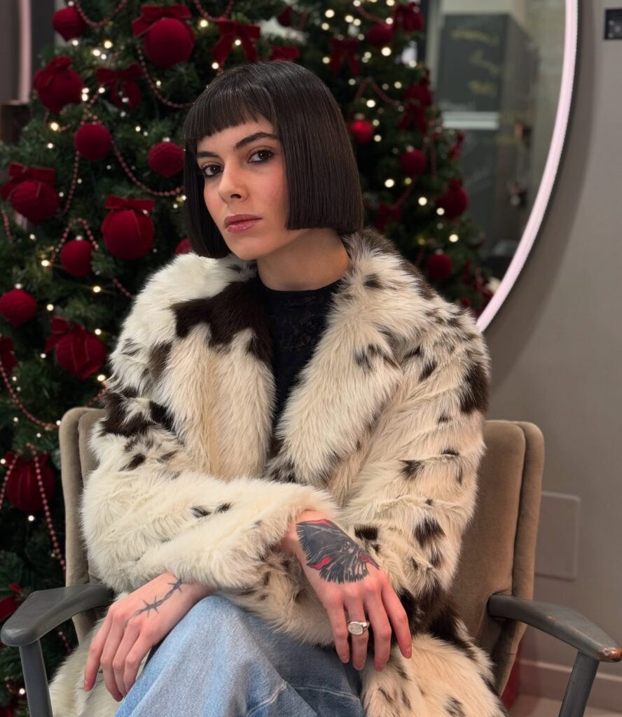 A woman sitting in a chair showcasing a very sharp, jaw-length dark brown blunt bob with short straight-cut micro bangs.