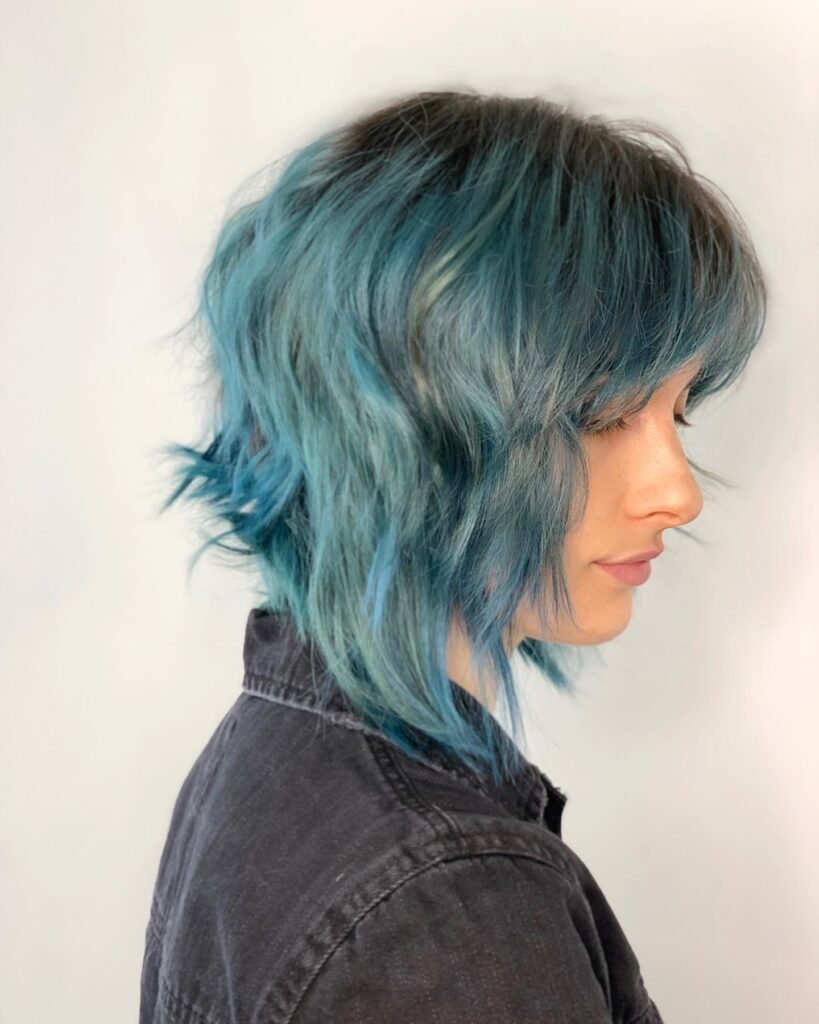 A woman in side profile showcases a textured, shaggy inverted bob with a dramatic teal blue color and dark roots