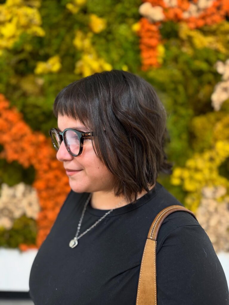 A woman in profile wearing glasses showcases a dark espresso choppy bob with a short arched micro fringe and soft textured waves.
