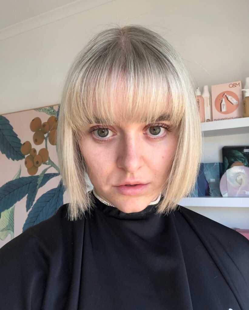 A woman looking directly forward showcases a straight ice blonde choppy bob with a full eyebrow-grazing fringe and subtle shadow roots.