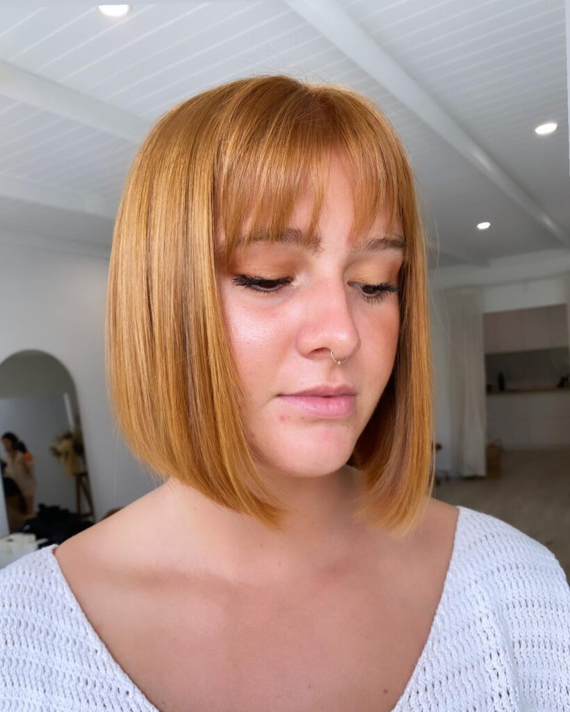 A close-up shot of a woman with a sleek, vibrant ginger-copper bob featuring blunt ends and straight-across fringe.