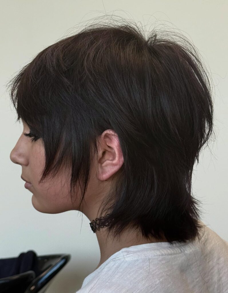 A woman in profile showing a textured dark brown shaggy bob hairstyle with short crown layers and wispy face-framing bangs.