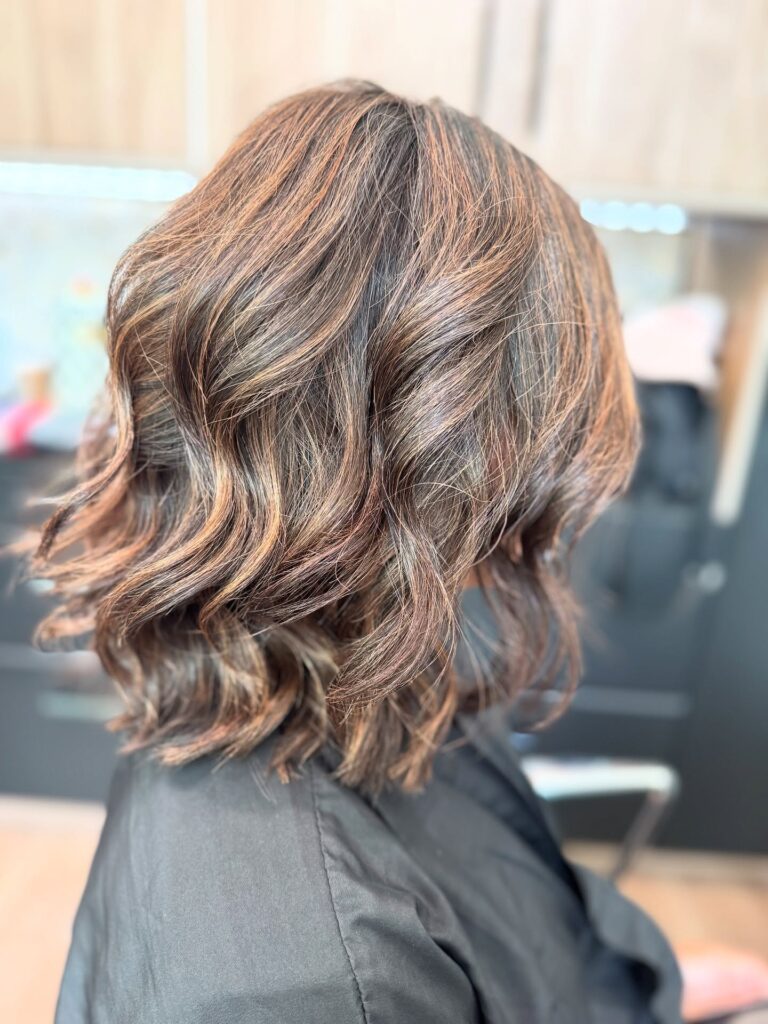A side profile showing a shoulder-length brunette bob haircut with textured waves and warm copper highlights.