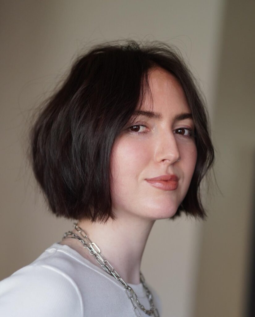 A woman looking toward the camera showcasing a voluminous chin length dark chocolate bob with a center part and textured waves.