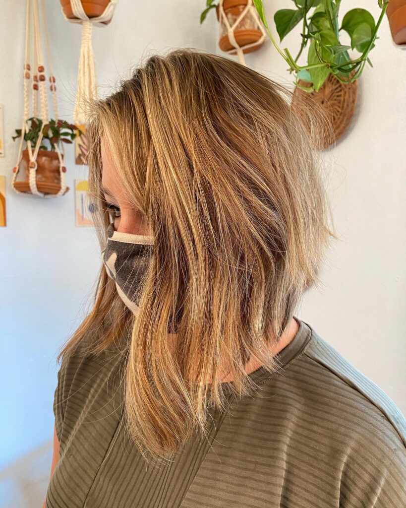 A woman from a side profile view showing a honey blonde angled A-line bob haircut with long side-swept bangs.