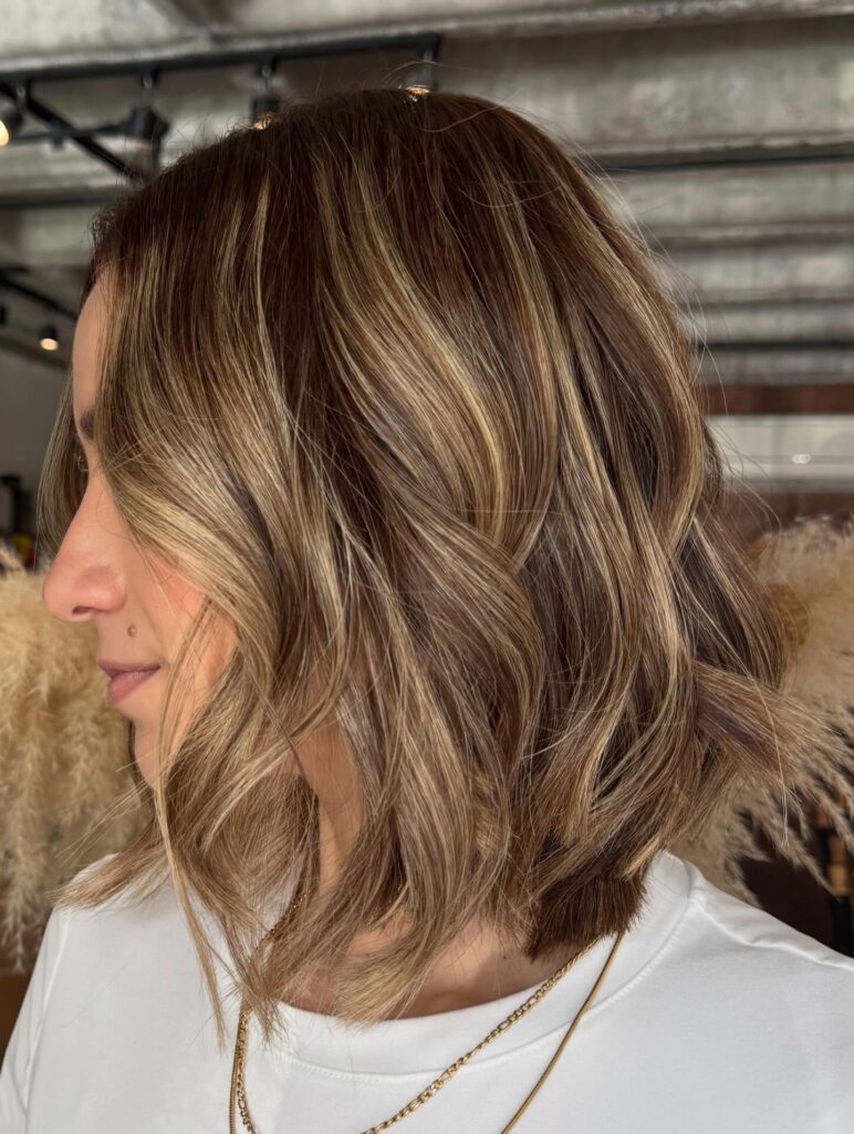 A woman in side profile showcasing a jaw length choppy honey blonde bob hairstyle with wavy layers and bright sun kissed highlights.