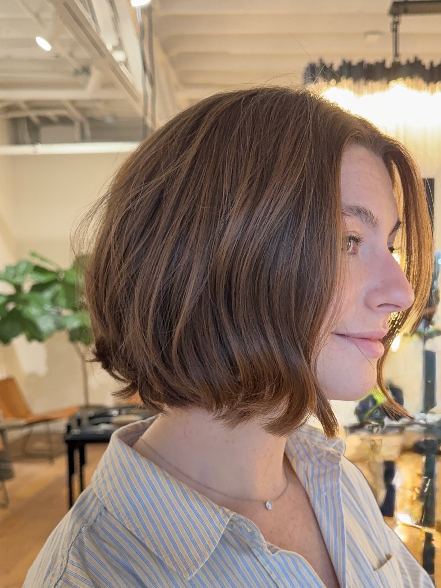 A woman smiling in side profile showcasing a jaw length choppy chestnut brown bob hairstyle with soft waves and a gentle lift at the back.