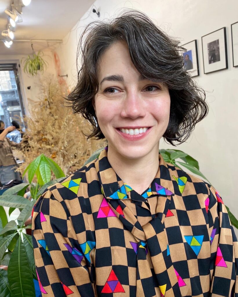 A smiling woman showcasing a jaw length choppy dark brown bob hairstyle with natural silver highlights and textured wavy layers.